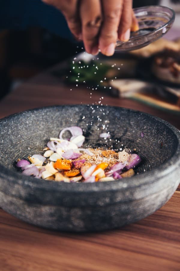 Salt falling from a hand onto sliced shallots and garlic in a stone mortar